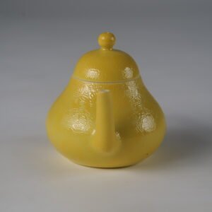 Eye-level front view facing the spout of a bright yellow pear-shaped porcelain teapot on white background