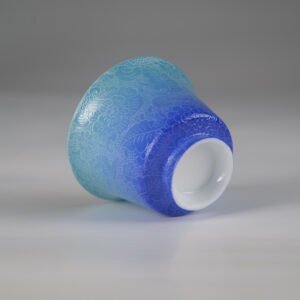 An angled bottom-up view of the East Breaking Dawn gradient blue porcelain horseshoe cup, showing the smooth white foot rim and full-body floral engraving