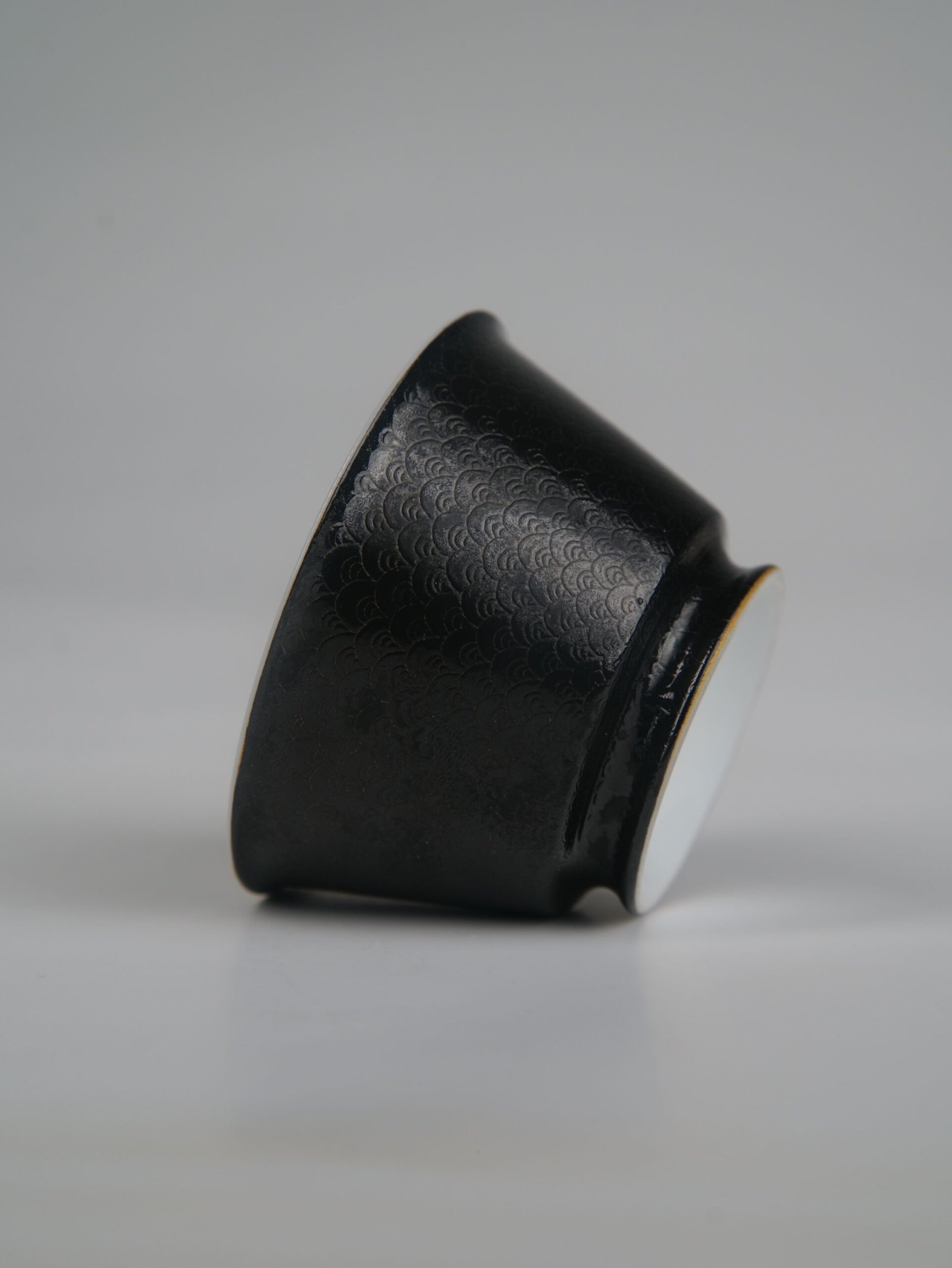 obsidian-black-linked-loop-porcelain-cup-side-profile-view Side profile view of the Obsidian Black Linked-loop porcelain cup lying on its side, highlighting its dark carved enamel dragon scale pattern and smooth base
