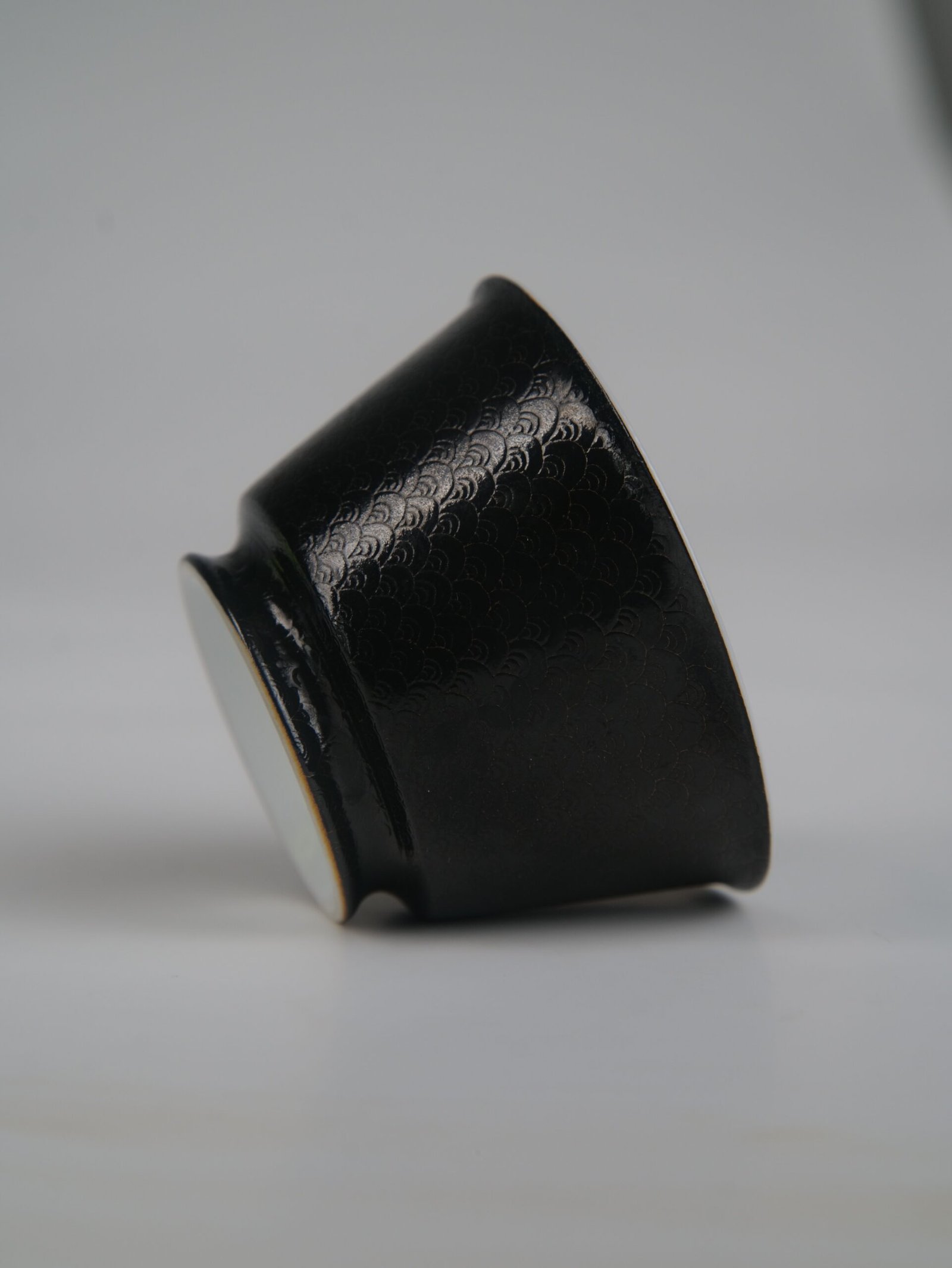 obsidian-black-linked-loop-porcelain-cup-tilted-side-view Tilted side view of the Obsidian Black Linked-loop porcelain cup lying on its side, showing its elegant curve, foot base, and carved dragon scale texture