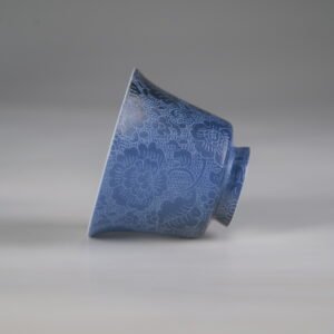 Horizontal side view of the Pale Ink Pool blue porcelain horseshoe cup lying on its side, showcasing its delicate curves and full-body floral carving