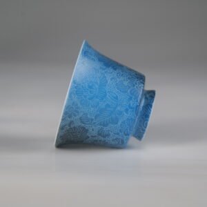 Side profile view of the Pale Ink Pool blue porcelain horseshoe cup lying on its side, showcasing its elegant shape and intricate floral carving