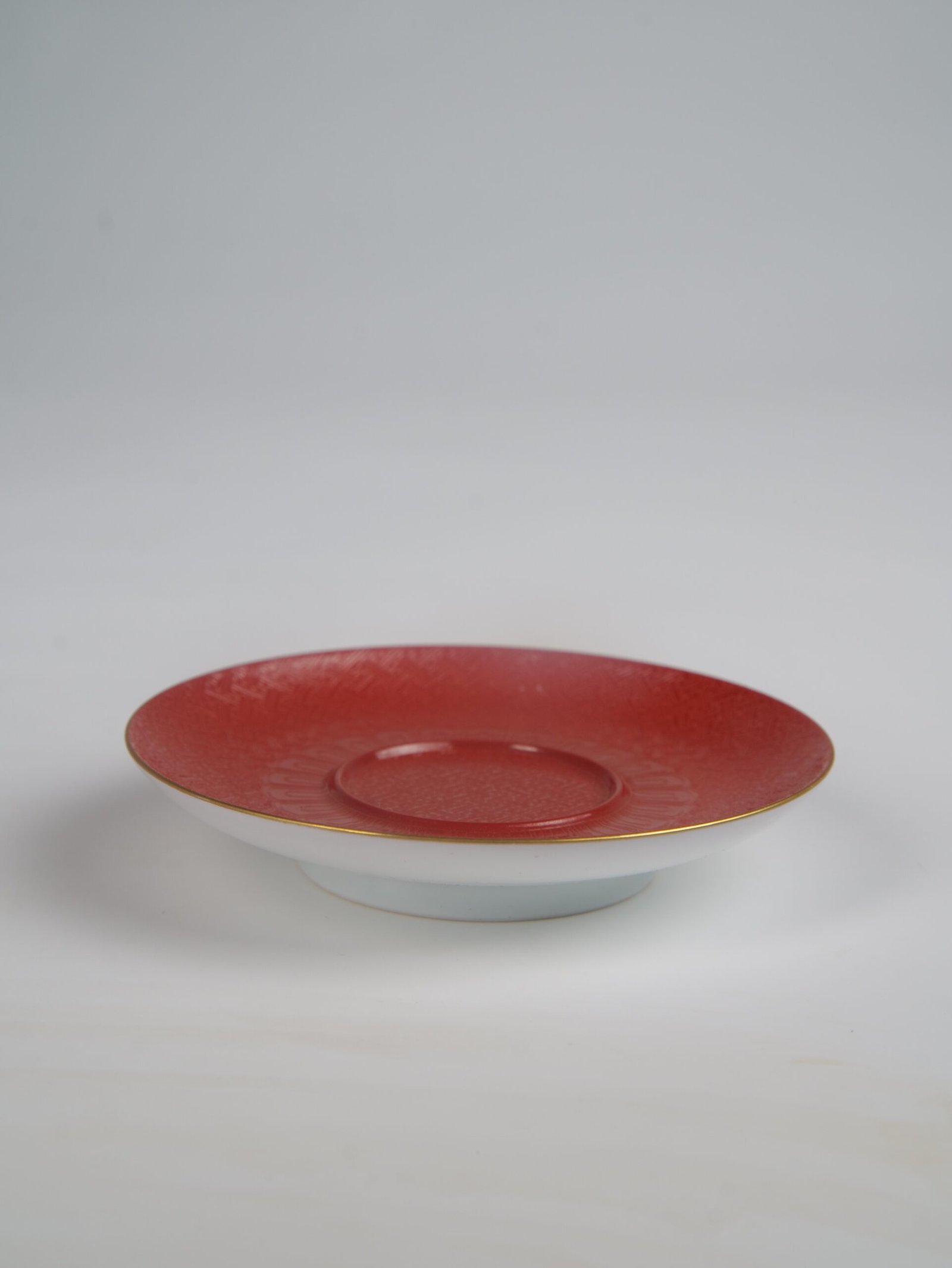 palace-red-elegant-charm-gaiwan-saucer The red porcelain saucer of the Palace Red Elegant Charm Gaiwan set with a gold-gilded rim and embossed pattern on a plain white background.