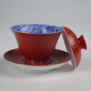 The Palace Red Elegant Charm Gaiwan with the lid removed, revealing the vibrant blue and white floral pattern lining the inside of the textured red cup, with the lid leaning against the saucer.