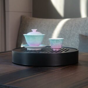 Lifestyle view of the Spring Glow Gradient Elegant Charm Gaiwan paired with a matching teacup on a black round tea tray in soft sunlight, showcasing the soft pink and fresh green gradient glaze and handcrafted Pa Hua pattern