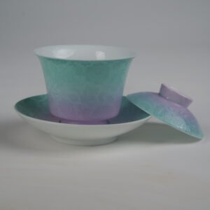 Side view of the Spring Glow Gradient Horseshoe Gaiwan with its lid removed and resting beside the saucer, showing the white porcelain interior, soft pink-purple and green gradient glaze, and fine engraved Pa Hua daylily patterns