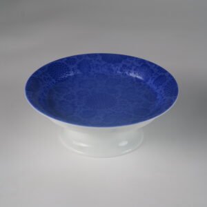 Angled view of ultramarine carved porcelain plate with white pedestal base